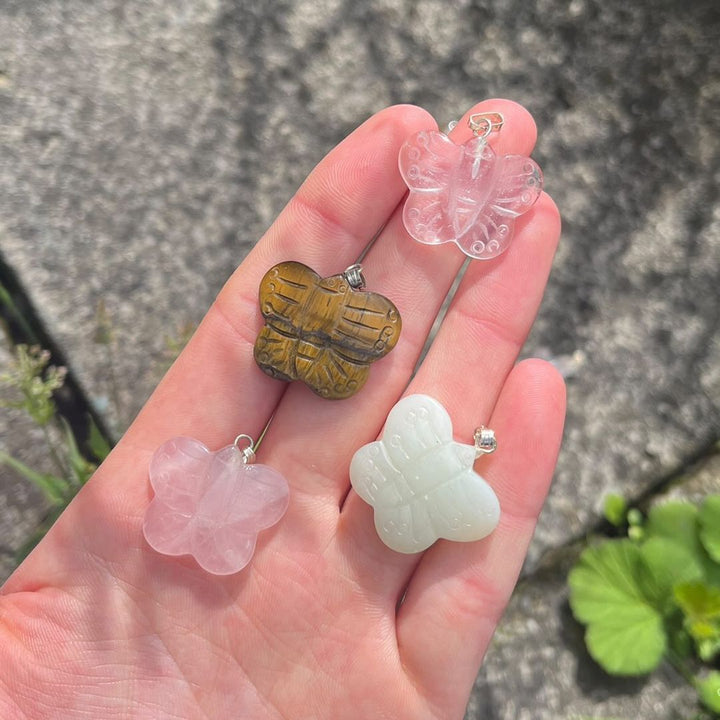 A close-up shot of a hand displaying small carved crystal pendants, showcasing a pink Rose Quartz butterfly and a golden brown Tiger Eye animal carving.