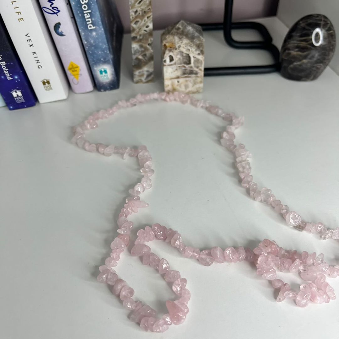 A long 32-inch rose quartz crystal chip necklace gracefully looped on a clean white table, showcasing polished, translucent pastel pink gemstone pieces styled indoors in front of books and decorative stone towers for Heart Chakra energy.