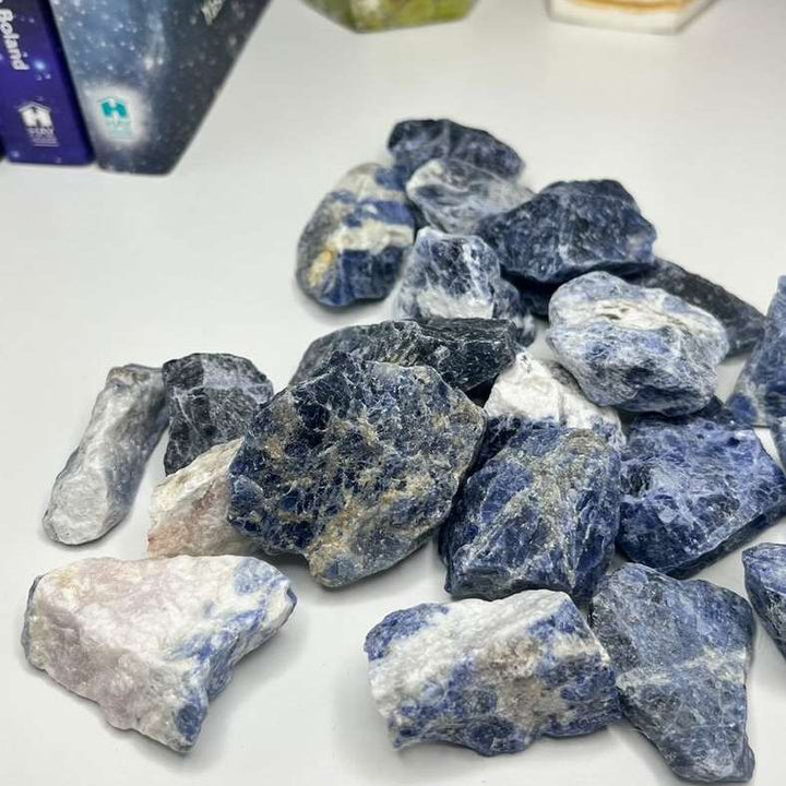 An eye-level shot looking down at a collection of raw blue sodalite crystal chunks spread across a smooth white surface. The crisp indoor lighting highlights their rugged angles, deep crevices, and natural denim tones, presenting a comforting and grounding presence for a bedside table or study desk.