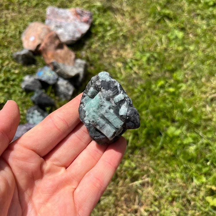 A close-up view of a hand presenting a single 3cm to 4cm rough emerald stone outdoors, showcasing the authentic deep green coloration and unique geological birthmarks left by the Earth to demonstrate its pocket sizing scale.