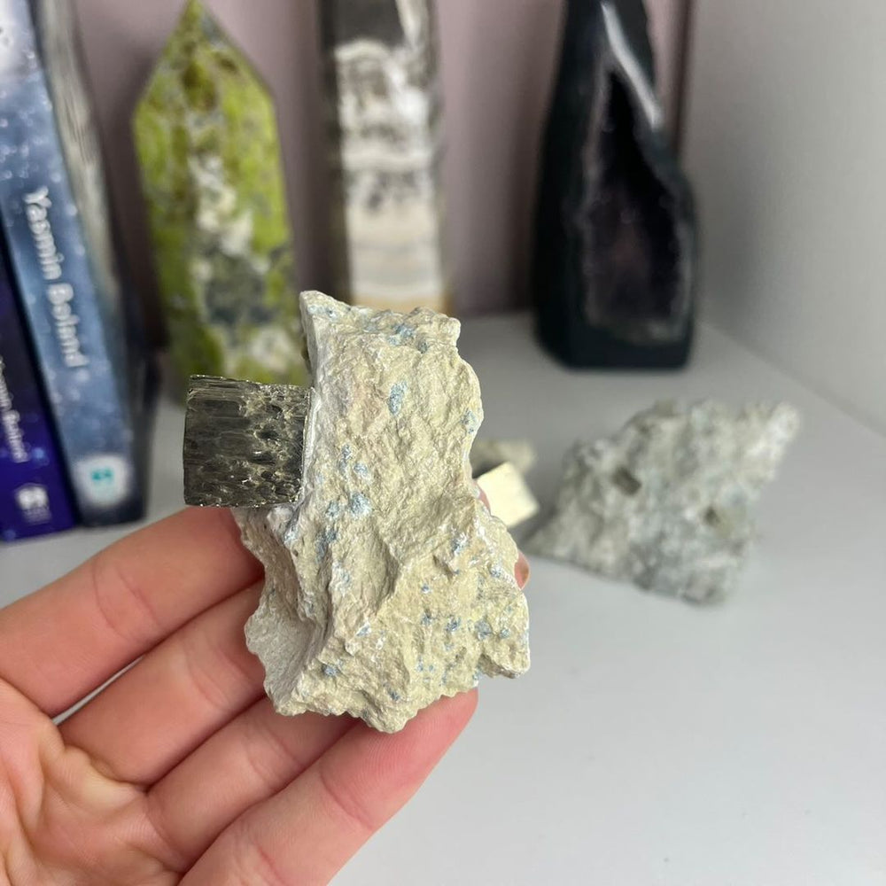A hand holding a single rough pyrite in matrix specimen vertically from the clearance collection, showing the stark unevenness and a prominent shimmering gold crystal pocket peeking out from the light grey host stone.