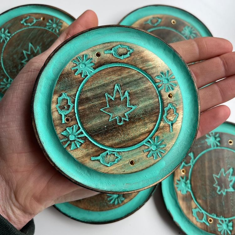 An open hand cradling a round turquoise wood incense burner plate outdoors, illustrating its compact 10cm width sizing scale, shallow curved rim, and small stick pre-drilled holes against a background of scattered matching ash catchers.

