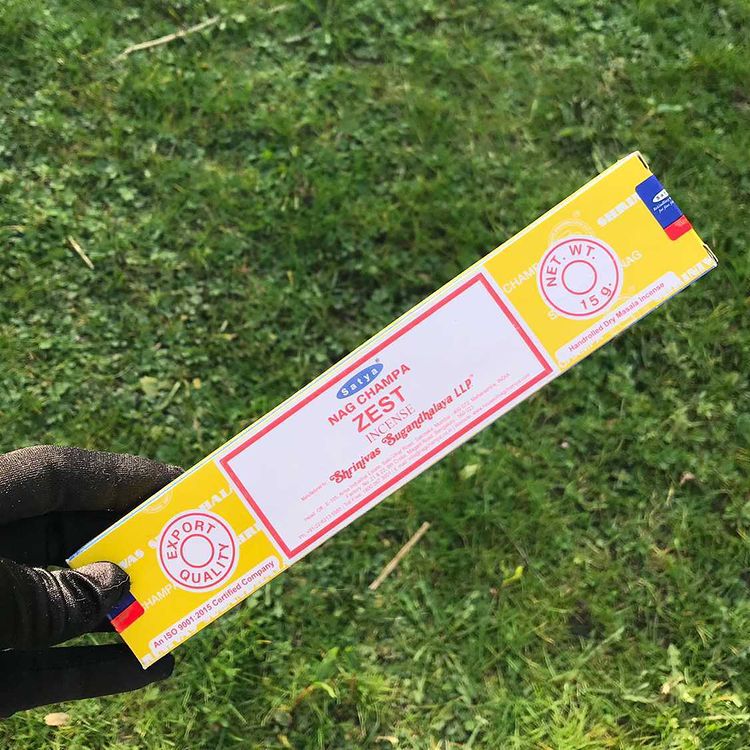 An overhead view looking down at a hand cradling a bright yellow and white rectangular box of Satya Zest incense sticks. The traditional hand-rolled packaging is displayed against a rich, sunlit background of green grass to mirror its sparkling citrus notes, awakening inner sparks and sunny Sacral chakra energy.