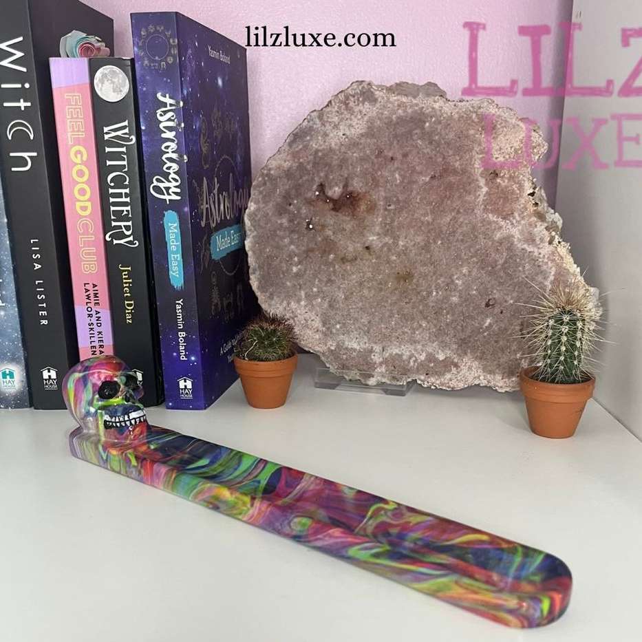 A close-up view of the long skull rainbow incense stick burner resting on a white tabletop shelf. The sleek boat-shaped tray features a detailed multi-coloured skull motif at one end, swirling with vibrant shades of violet, turquoise, yellow, and magenta.

