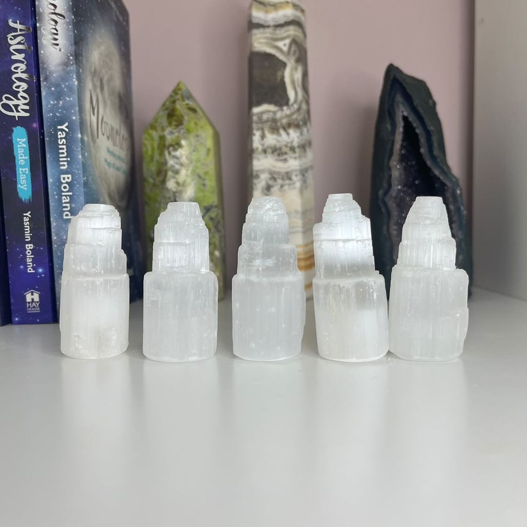 A row of five polished small selenite towers standing upright on a white altar shelf, showcasing their ice-white vertical columns, fibrous mineral layers, and chiselled points styled indoors in front of spiritual books and crystal towers for Crown Chakra alignment.