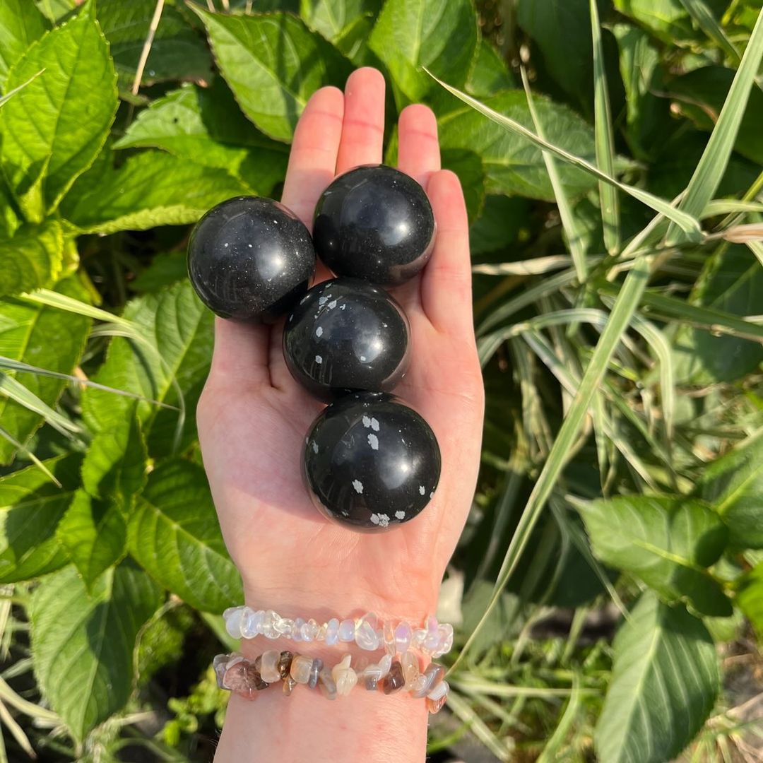 An open hand wearing stacked crystal chip bracelets holding four polished snowflake obsidian crystal spheres outdoors, showcasing their smooth round shape, deep black volcanic glass base, and unique white cristobalite patches against a lush background of garden leaves for Root Chakra grounding.