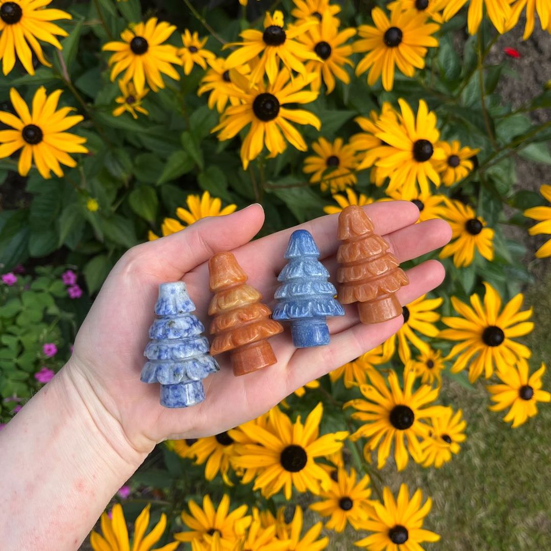 An open hand holding four hand-carved crystal trees outdoors, displaying two deep blue sodalite trees with white calcite streaks and two warm peach aventurine trees styled against a vibrant backdrop of blooming yellow flowers for Throat Chakra balancing.