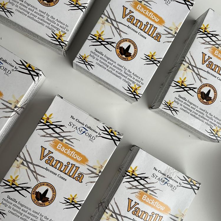 A top-down, flat-lay view of multiple rectangular boxes of Stamford Backflow Vanilla Incense Cones scattered across a clean, minimalist off-white surface. The crisp white packaging features gold text accents and illustrations of delicate orchid blossoms, highlighting the traditional hand-rolled quality designed to release a rich, creamy vanilla aroma for a relaxing everyday vibe.