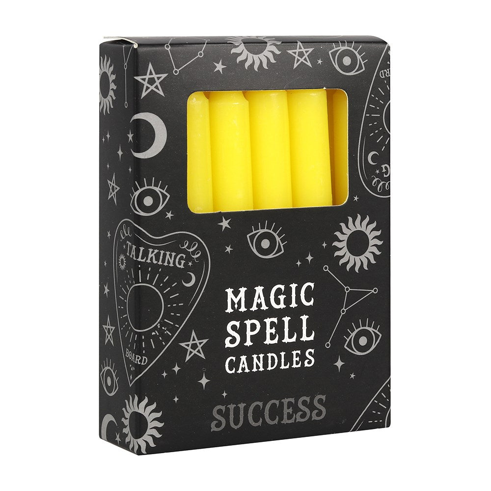 A three-quarter view of a black cardboard pack of Success spell candles against a white background. The box displays mystical white line illustrations of stars, crescent moons, and a talking board planchette, with a clear rectangular cutout window revealing several smooth, vibrant yellow unscented mini tapers designed to align with the Heart chakra.