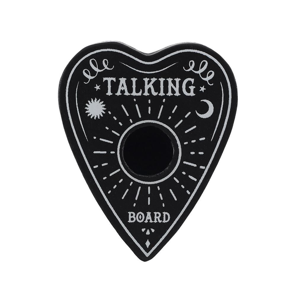 A flat-lay view of a black heart-shaped spell candle holder against a plain white setting. Shaped like a mystical talking board planchette, it displays crisp white engravings of celestial sun and moon illustrations alongside decorative line work to channel a thoughtful Third Eye chakra routine.