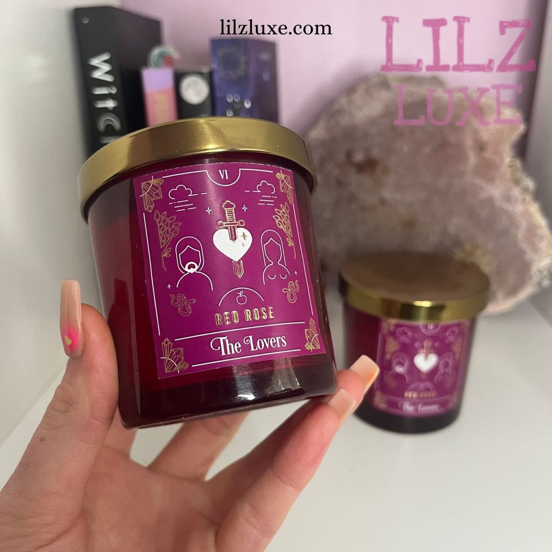 A hand with ombré pink nails presenting a red rose scented Lovers tarot card candle in a deep red glass jar with a metallic gold lid, styled indoors next to a large raw pink quartz cluster and spiritual books for Heart Chakra alignment.