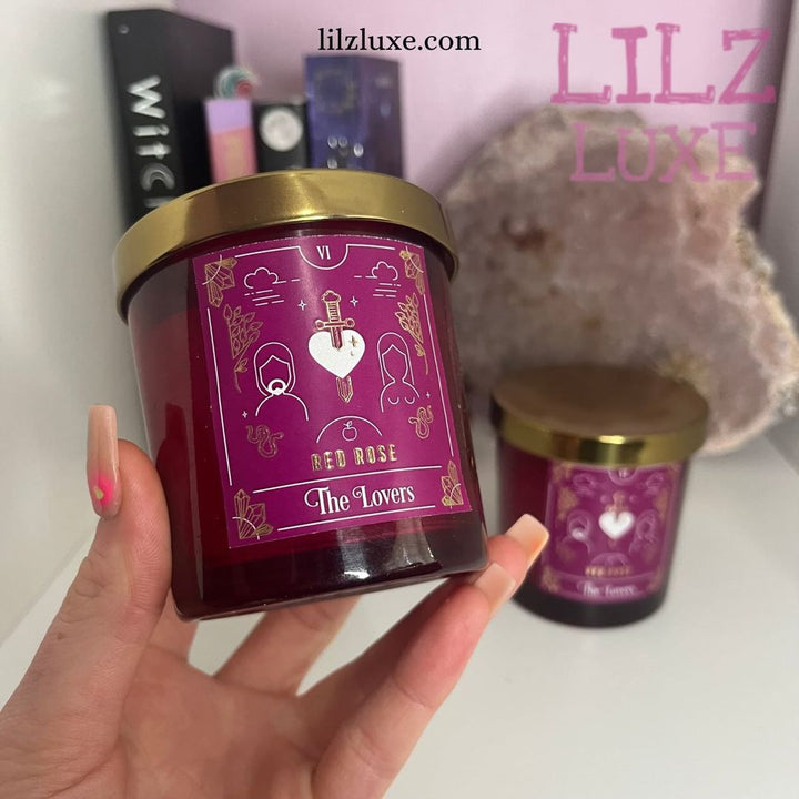 A hand with ombré pink nails presenting a red rose scented Lovers tarot card candle in a deep red glass jar with a metallic gold lid, styled indoors next to a large raw pink quartz cluster and spiritual books for Heart Chakra alignment.