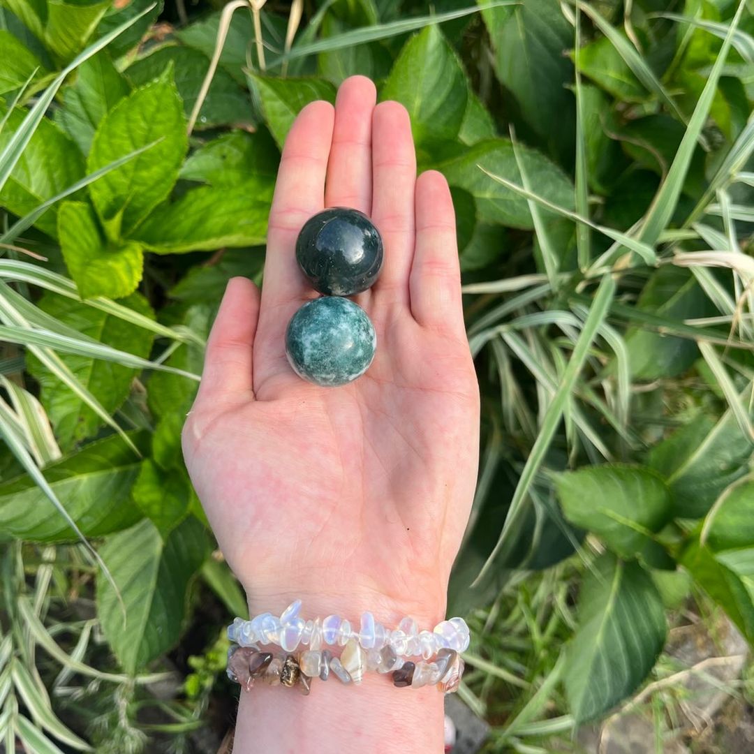 An open hand wearing stacked crystal chip bracelets holding two polished tree agate spheres outdoors, showcasing a striking canvas of forest green branching patterns against a snowy white backdrop and a compact 3cm sizing scale against a fresh background of green garden foliage.
