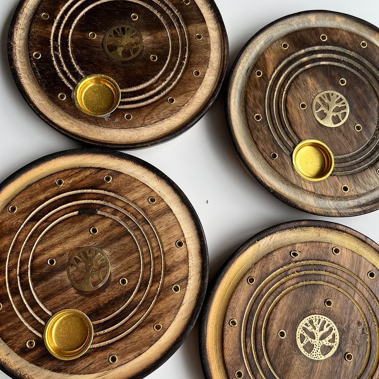 An overhead shot of four round, dark mango wood incense holders designed for sticks and cones, showcasing concentric ring grooves, multiple pre-drilled stick placement holes, a small brass cup for burning incense cones safely, and a carved Tree of Life emblem in the centre.