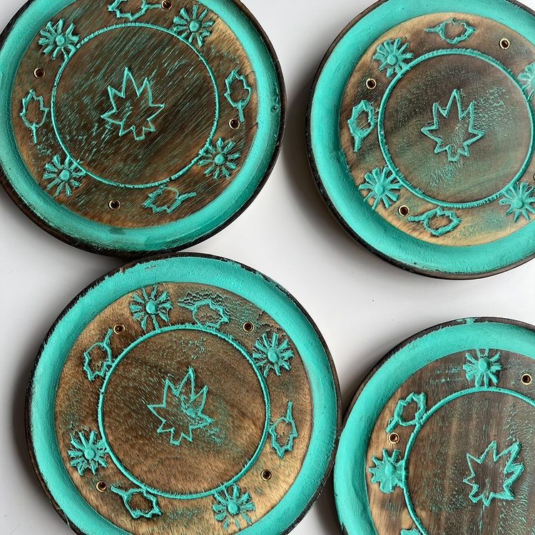 A flat lay shot displaying four round turquoise incense stick holders, showcasing the raw mango wood grain centers detailed with carved celestial stars and leaf patterns outlined in vibrant painted turquoise tones to create a serene sanctuary vibe.
