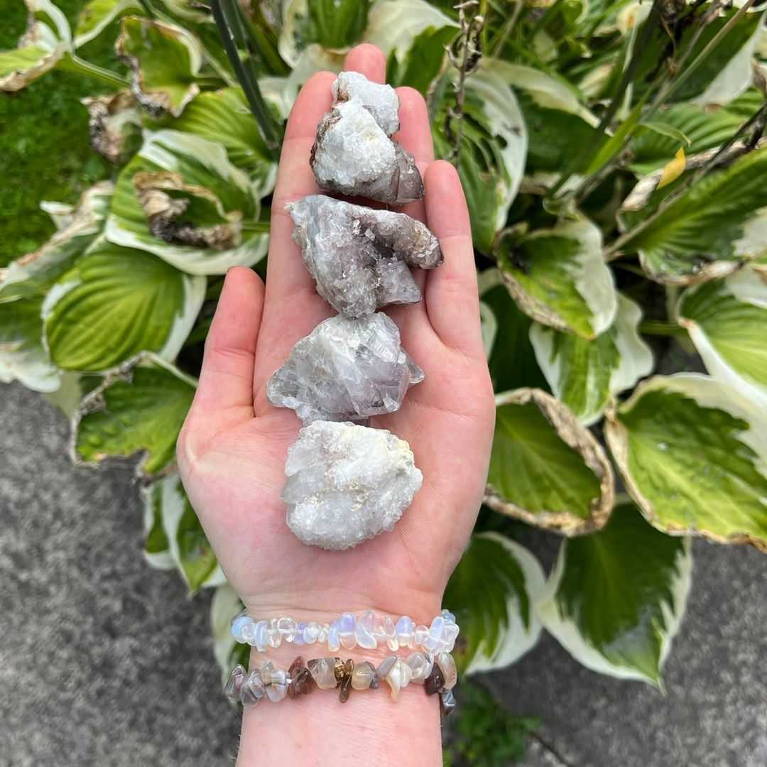 A straight vertical view of a hand supporting a stack of raw, light purple Spirit Quartz cluster specimens. The natural framing emphasizes the crystalline textures and authentic earthy qualities of these unique pocket treasures against a rich background of garden foliage.