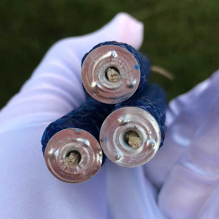 An angled bottom-up shot focusing on the flat circular bases of three dark blue beeswax manifestation candles resting inside an open, white gloved hand. This detail shot highlights the small metal wick tabs at the core and the uniform thickness of the hand-rolled sheets used for setting peace intentions.