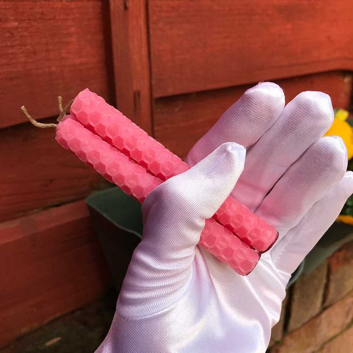 A side profile view of three texturised pink beeswax ritual candles held horizontally across a white gloved palm. The layout showcases the sweet pink colour palette, clean edges, and detailed honeycomb pattern of the taper set against a soft-focus natural garden backdrop.